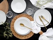 Draufsicht auf ein elegant gedecktes Tischarrangement mit weißen, geriffelten Porzellantellern und -schalen, goldfarbenem Besteck, geflochtenem Platzset, Glasuntersetzern und weißer Serviette auf dunklem Untergrund.