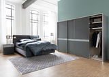 Modernes Schlafzimmer mit einem grauen Polsterbett, zwei Nachttischen und einem großen Kleiderschrank mit Schiebetüren. Perspektive von vorne links.