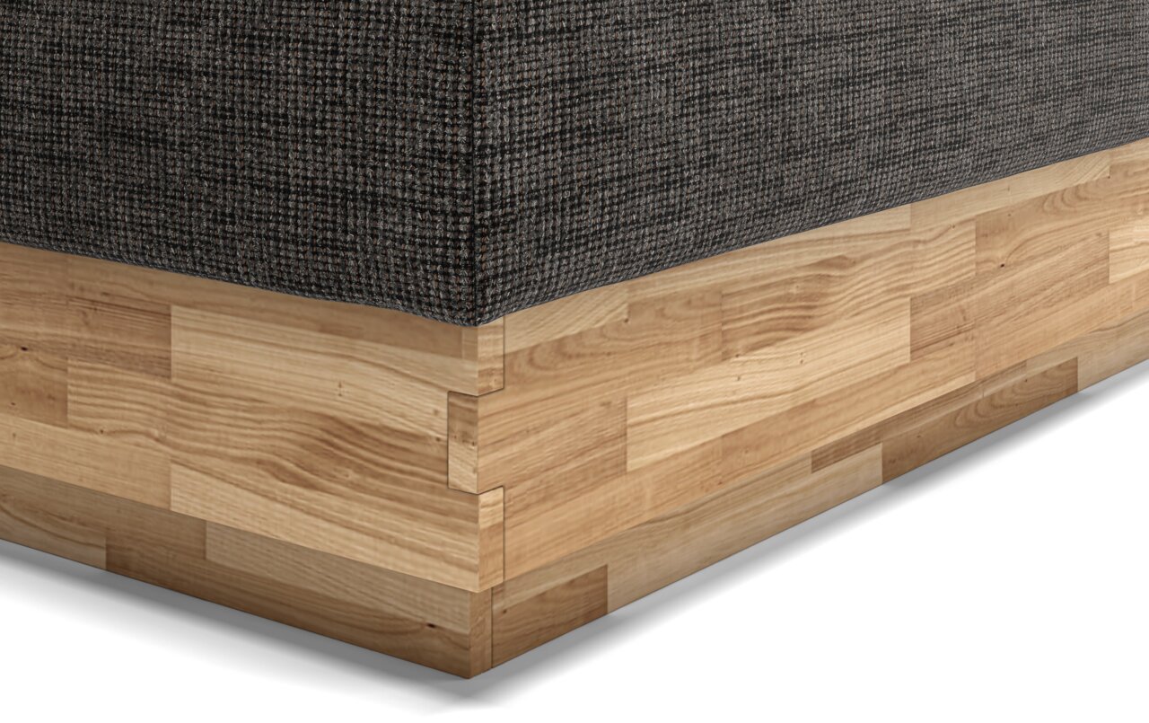 Nahaufnahme eines Boxspringbettes mit dunklem Stoffbezug und hellem Holzunterbau, perspektivisch von der Ecke aufgenommen.