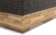 Nahaufnahme eines Boxspringbettes mit dunklem Stoffbezug und hellem Holzunterbau, perspektivisch von der Ecke aufgenommen.