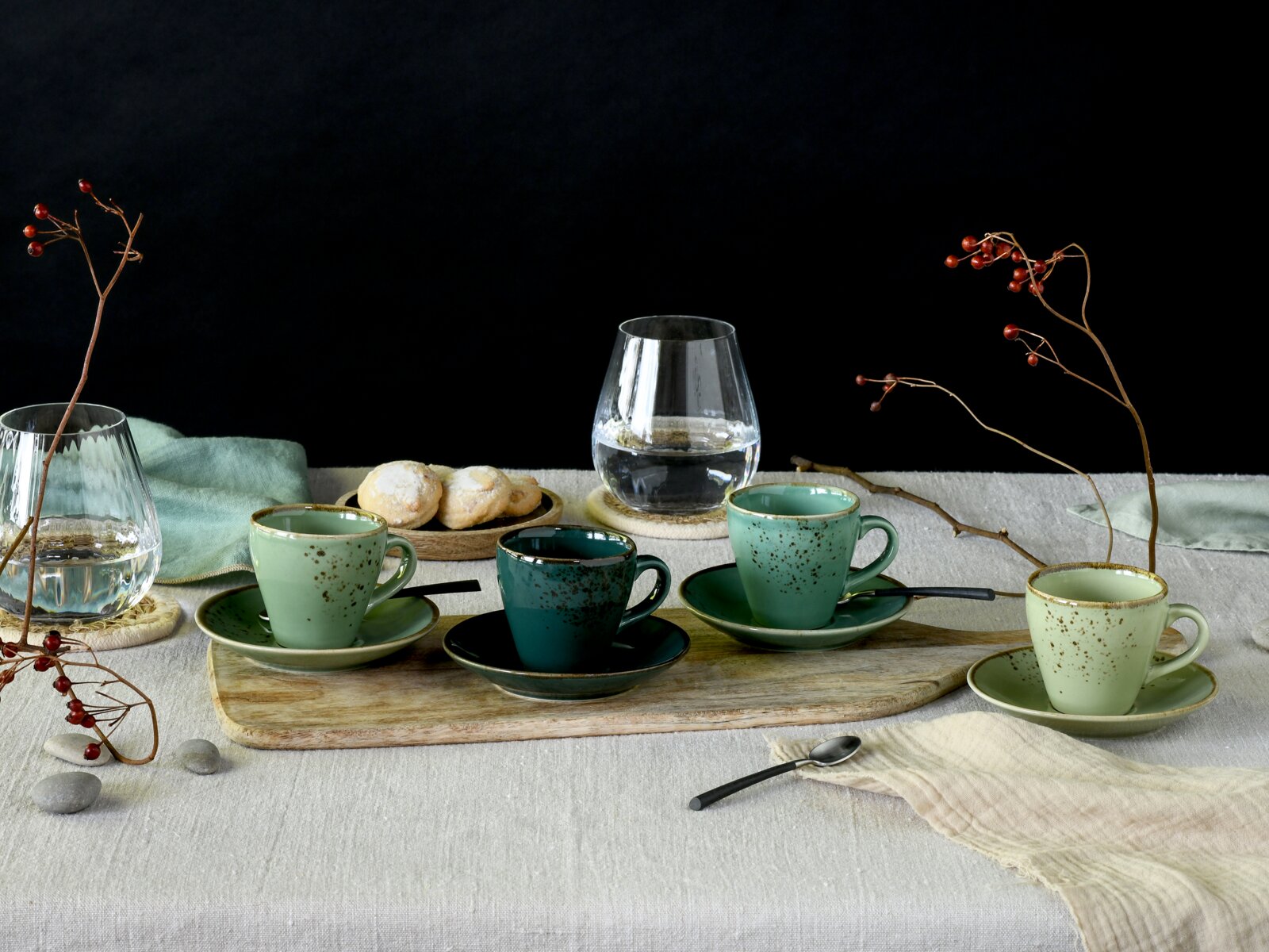Rustikales Kaffee-Set mit vier gesprenkelten Keramik-Tassen und Untertassen in Grün- und Blautönen auf Holzbrett, dazu Glaskaraffe, Kekse und Beerenzweige auf Leinentuch vor schwarzem Hintergrund, Perspektive: leicht erhöhte Frontansicht