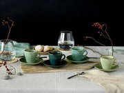 Rustikales Kaffee-Set mit vier gesprenkelten Keramik-Tassen und Untertassen in Grün- und Blautönen auf Holzbrett, dazu Glaskaraffe, Kekse und Beerenzweige auf Leinentuch vor schwarzem Hintergrund, Perspektive: leicht erhöhte Frontansicht
