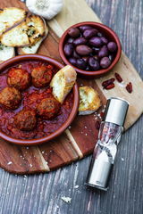 Gewürzschneider und Mühle DUOS aus Edelstahl, liegend auf einem rustikalen Holztisch neben einer Schale mit Fleischbällchen in Tomatensauce und einer Schale mit Oliven.