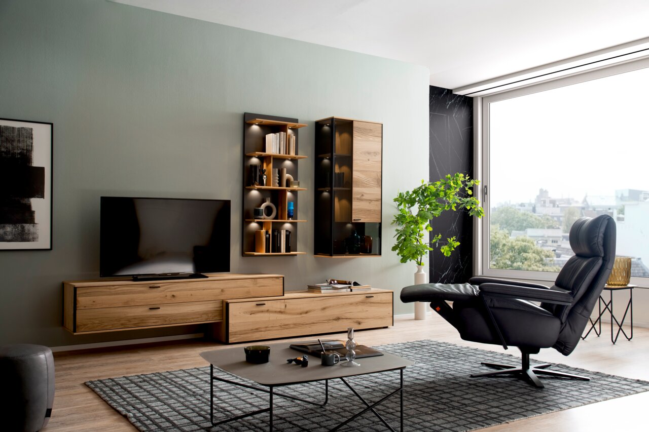 Moderner, drehbarer Relaxsessel in schwarzem Leder, seitliche Perspektive, in einem stilvoll eingerichteten Wohnzimmer mit großen Fenstern und Holzmöbeln.