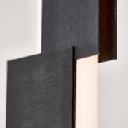 Detailansicht aus seitlicher Nahperspektive einer modernen Leuchte mit überlappenden, dunkel gebürsteten Metallflächen und warm leuchtendem Diffusor