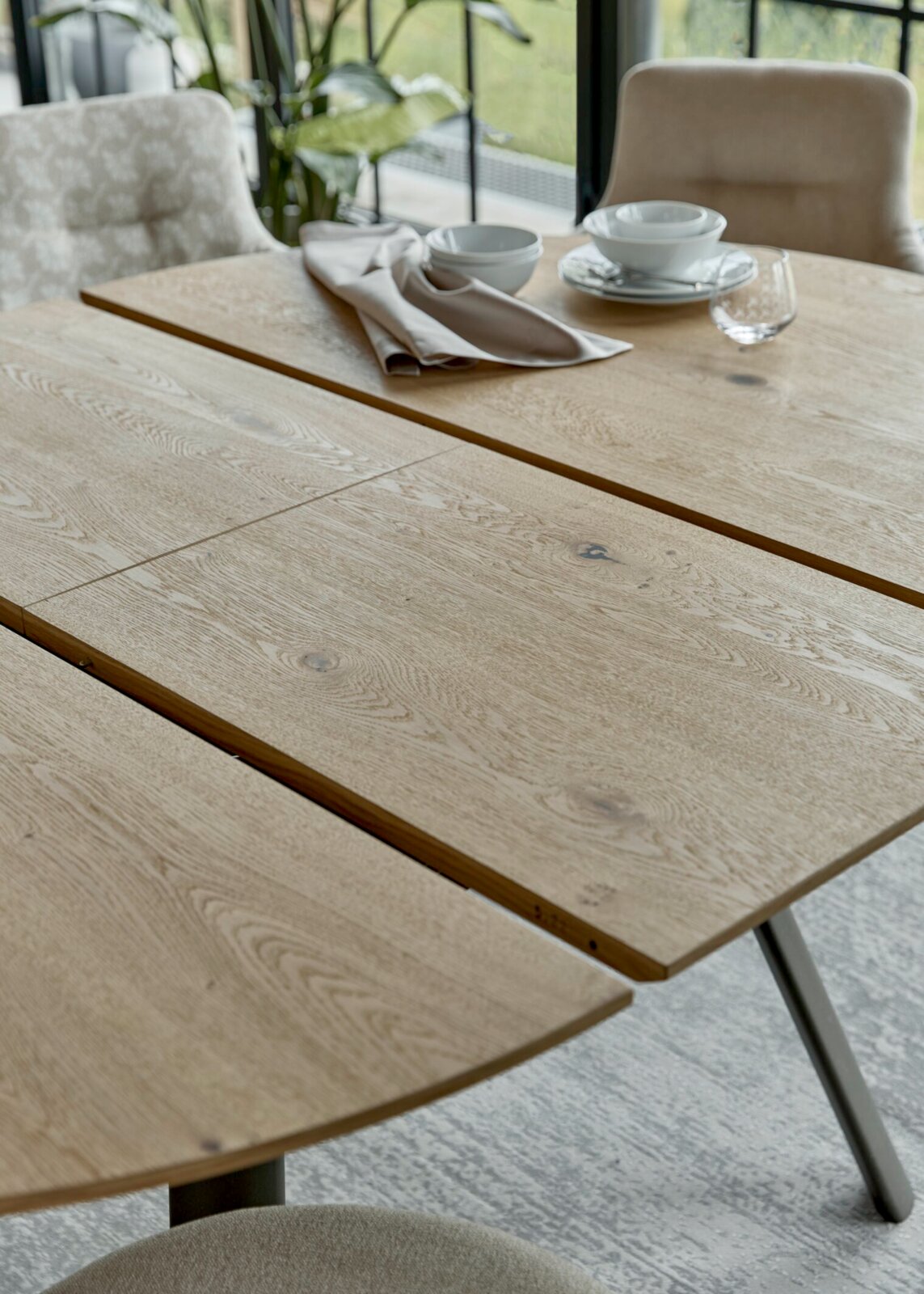 Ausziehbarer Esstisch aus hellem Holz mit sichtbarer Maserung und Metallbeinen, schräg von oben aus der Nähe fotografiert, mit Geschirr und Stoffserviette auf der Tischplatte.