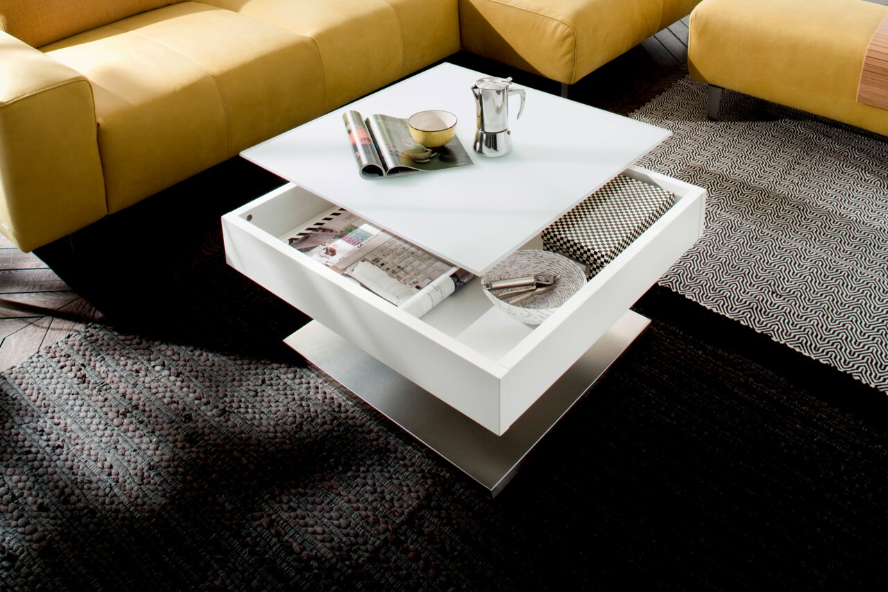 Moderne witte salontafel met lade, gefotografeerd van bovenaf, omringd door gele banken en tapijten.