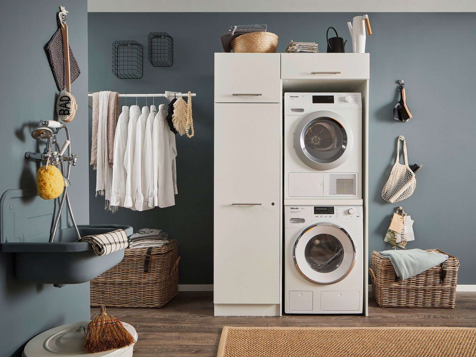 Hauswirtschaftsraum mit Waschmaschine und Trockner in einem weißen Hochschrank, umgeben von Körben und Hängeregalen, seitliche Perspektive.