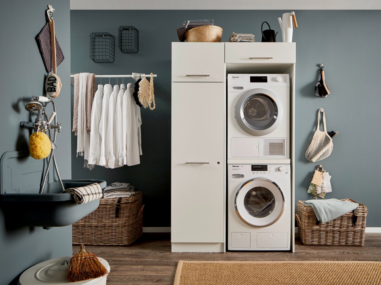 Hauswirtschaftsraum mit Waschmaschine und Trockner in einem weißen Hochschrank, umgeben von Körben und Hängeregalen, seitliche Perspektive.