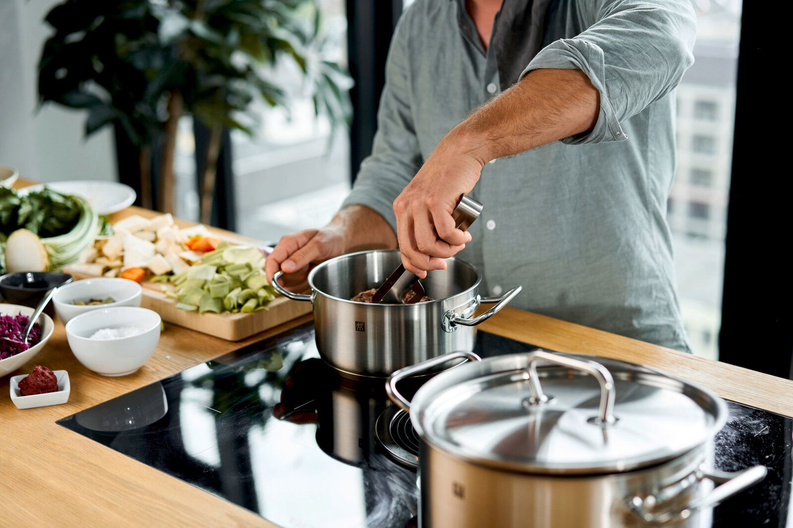ZWILLING Topfset 5-tlg. TWIN CLASSIC Mann kocht mit einem Edelstahl-Topf aus dem 5-teiligen Twin Classic Topfset auf einem Herd, mit Gemüse im Hintergrund. Perspektive: seitlich.