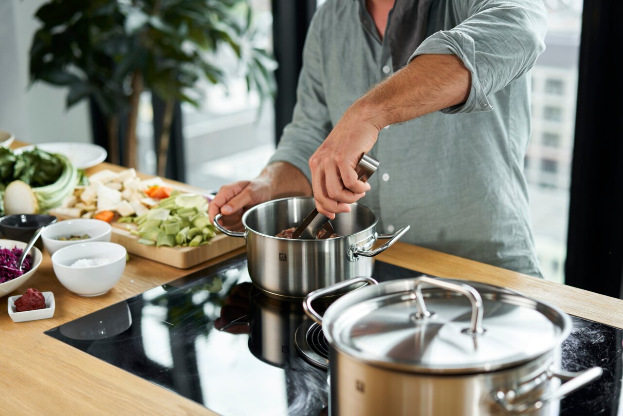Mann kocht mit einem Edelstahl-Topf aus dem 5-teiligen Twin Classic Topfset auf einem Herd, mit Gemüse im Hintergrund. Perspektive: seitlich.