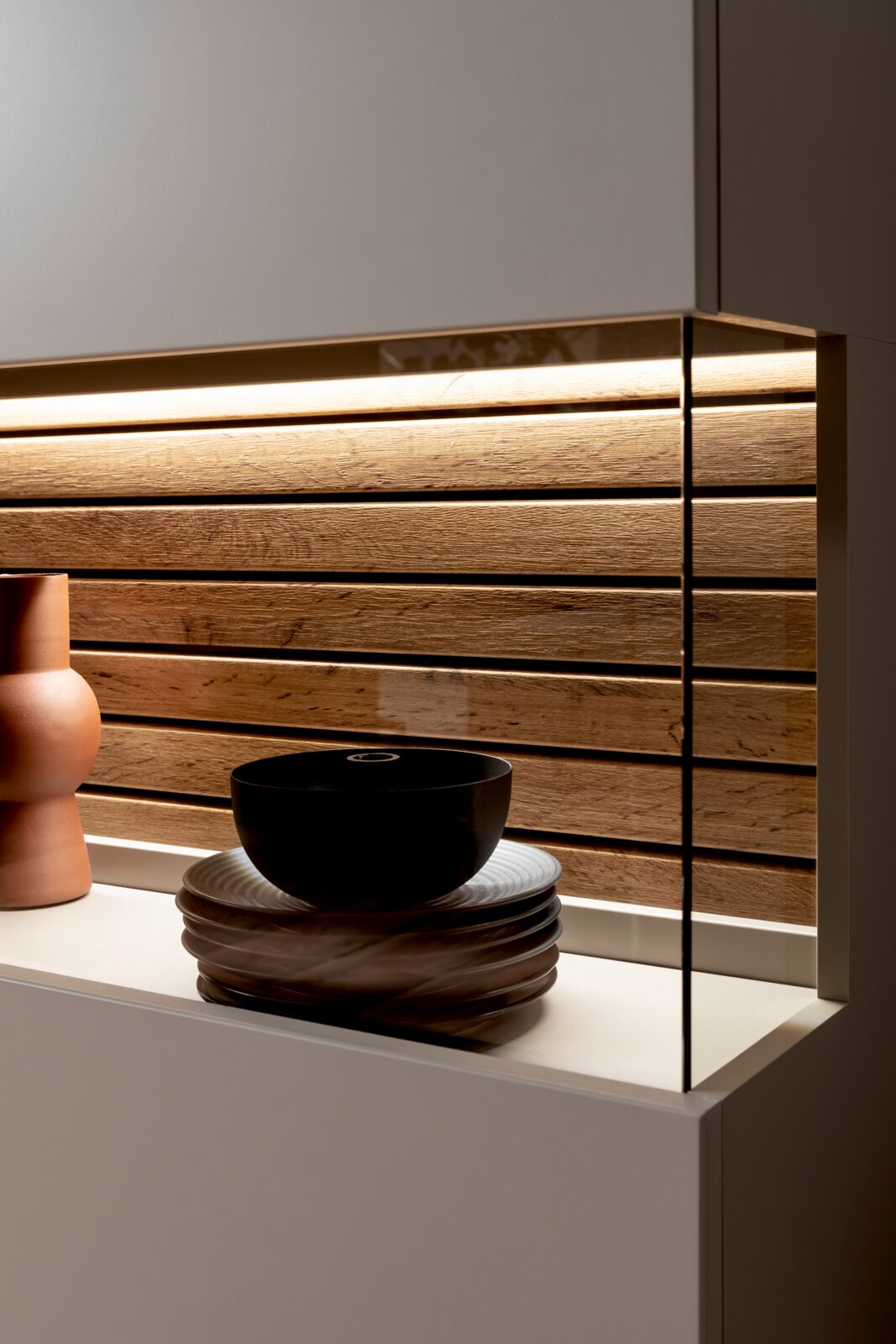 Moderne Küchenrückwand aus horizontalen Holzlamellen mit warmem LED-Licht, rechts mit Glas-Seitenpanel, Vorderansicht leicht von links auf Arbeitsplatte mit Geschirr und Vase