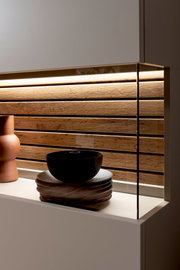 Moderne Küchenrückwand aus horizontalen Holzlamellen mit warmem LED-Licht, rechts mit Glas-Seitenpanel, Vorderansicht leicht von links auf Arbeitsplatte mit Geschirr und Vase
