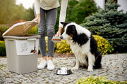 Tierfutterbehälter 38 l mit geöffnetem Deckel, eine Person füllt Hundefutter in einen Napf, während ein Hund daneben sitzt. Perspektive von vorne.