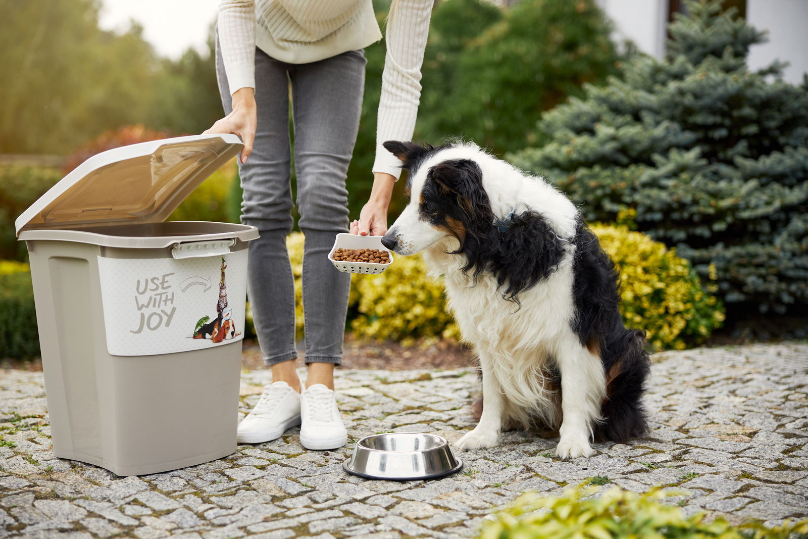 Tierfutterbehälter 38 l mit geöffnetem Deckel, eine Person füllt Hundefutter in einen Napf, während ein Hund daneben sitzt. Perspektive von vorne.
