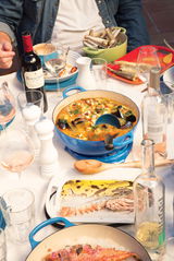 Ein gedeckter Tisch mit einer blauen Gusseisenpfanne voller Meeresfrüchte-Eintopf, umgeben von Weingläsern, Weinflaschen und verschiedenen Schalen mit Meeresfrüchten. Draufsicht.