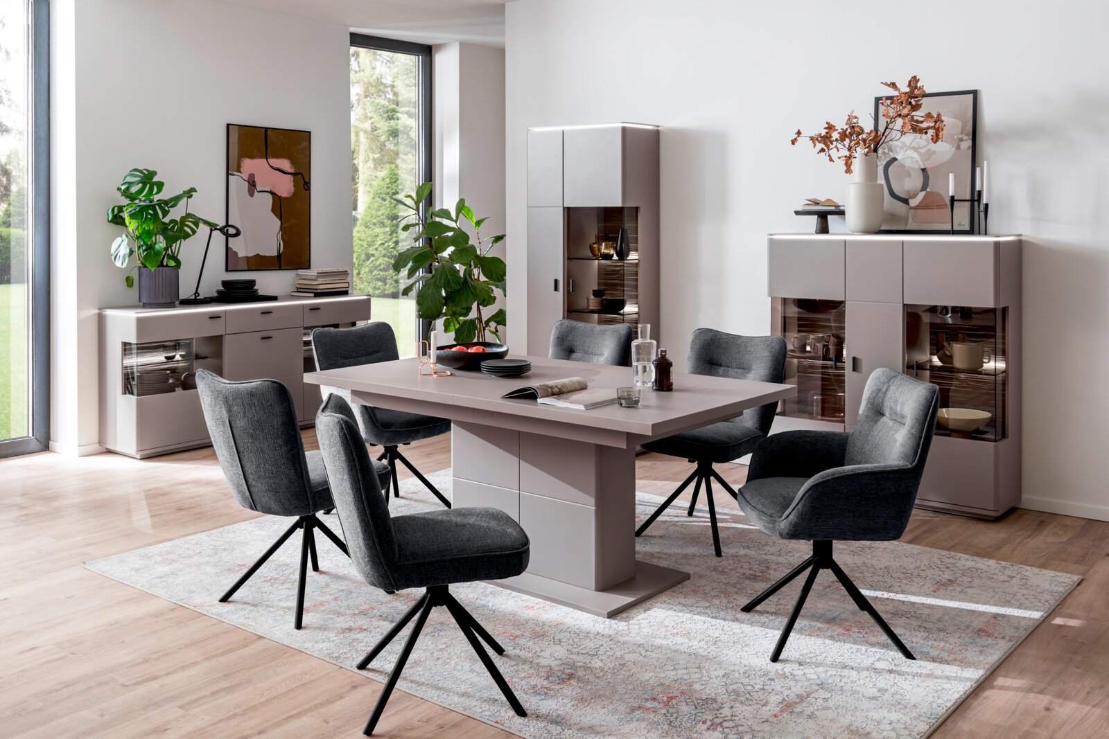 Modernes Esszimmer-Set mit rechteckigem Esstisch in Taupe/Grau und sechs gepolsterten, dunkelgrauen Drehstühlen auf gemustertem Teppich, umgeben von Sideboard und Vitrine – Perspektive: leicht erhöhte Frontansicht schräg von links.