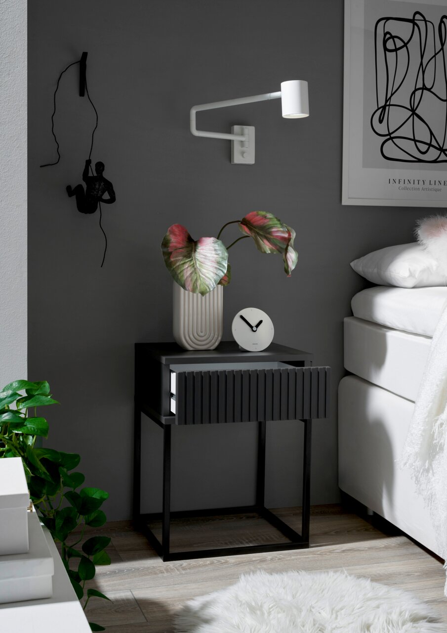 Moderne Nachtkommode in dunklem Design mit geriffelter Schubladenfront, dekoriert mit einer Pflanze und einer Uhr, seitliche Perspektive
