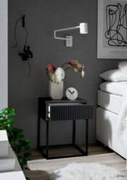 Moderne Nachtkommode in dunklem Design mit geriffelter Schubladenfront, dekoriert mit einer Pflanze und einer Uhr, seitliche Perspektive