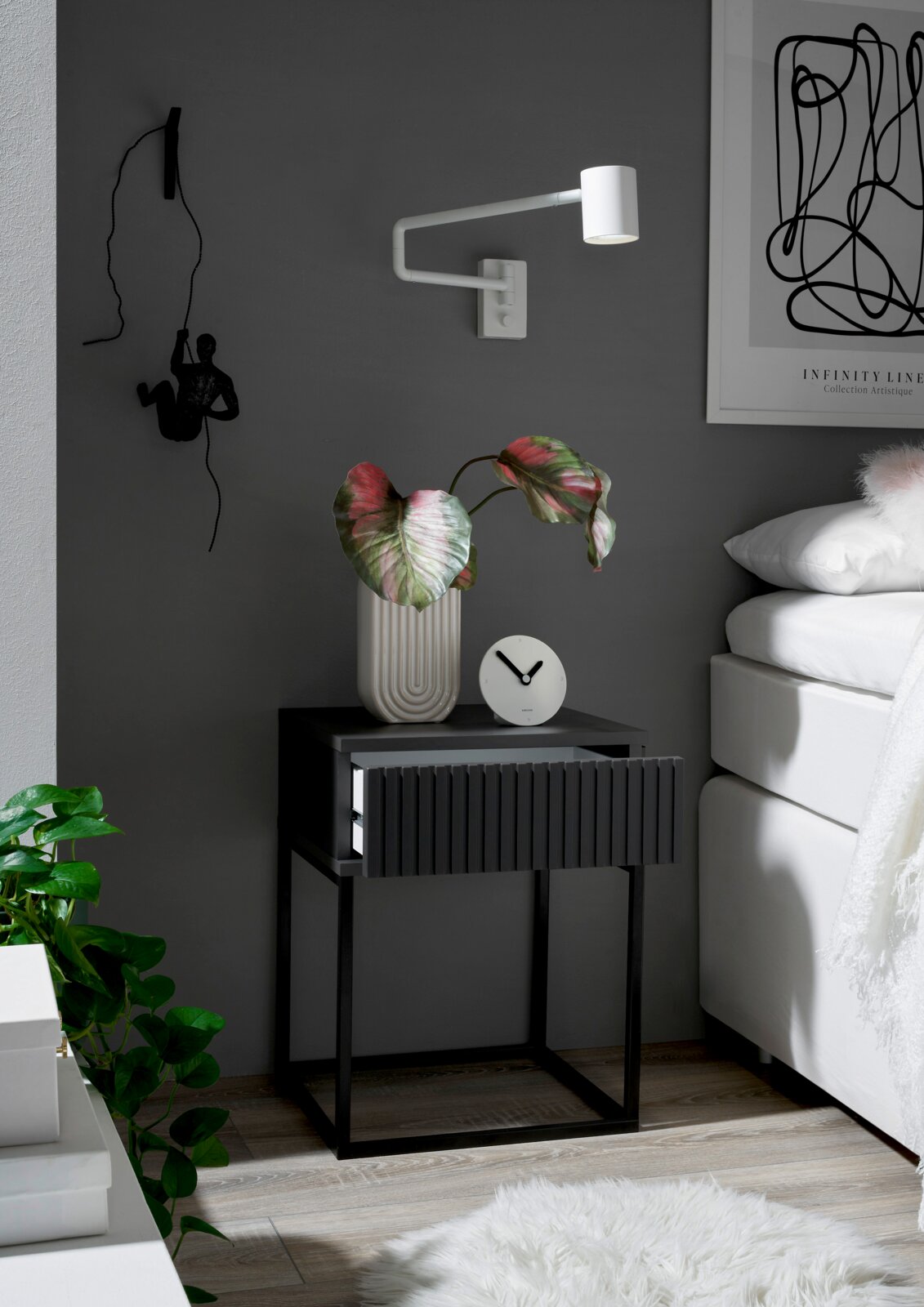 Moderne Nachtkommode in dunklem Design mit geriffelter Schubladenfront, dekoriert mit einer Pflanze und einer Uhr, seitliche Perspektive