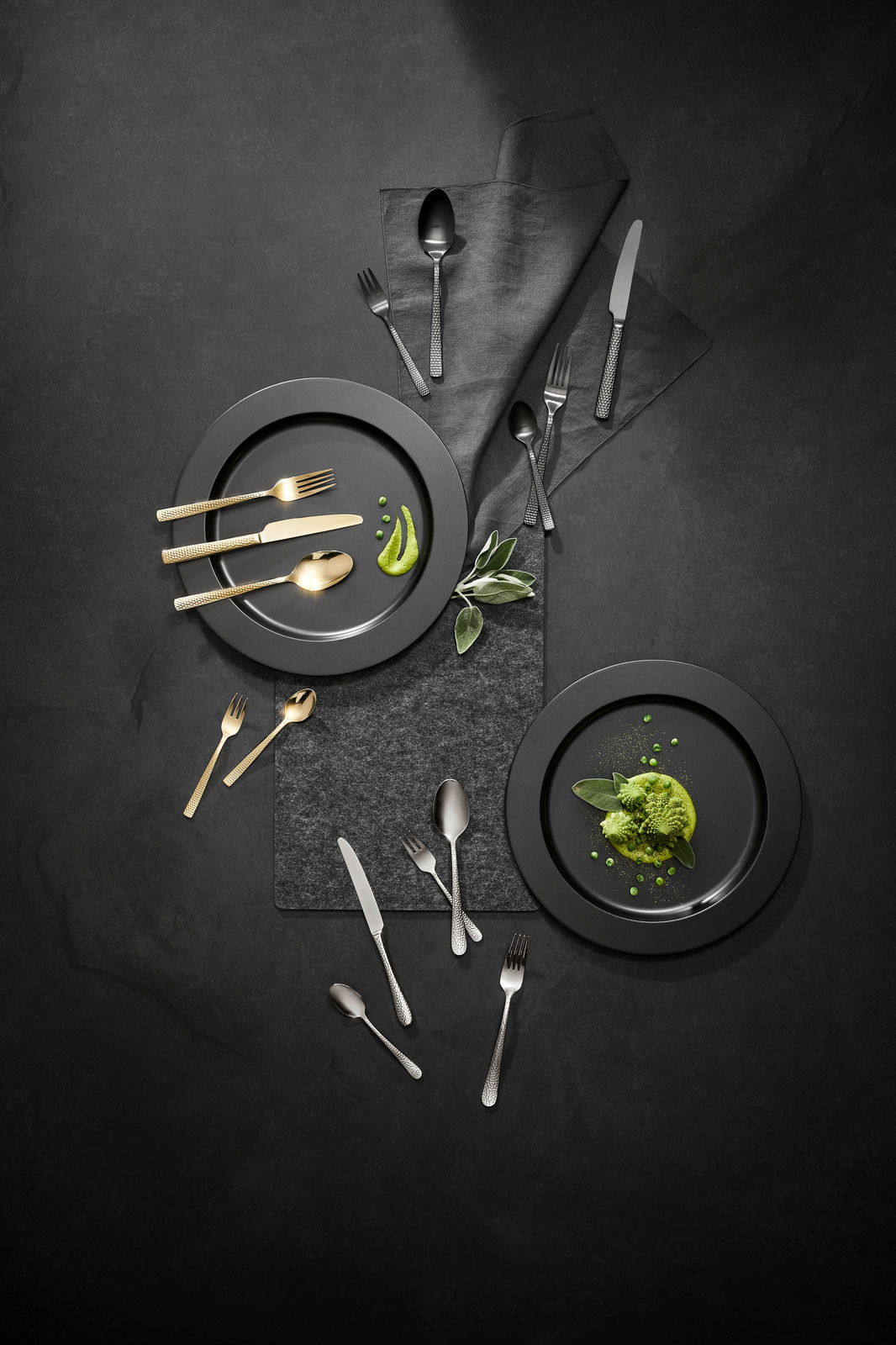 Vogelperspektive auf ein stilvolles Besteckset mit goldenen und silbernen Besteckteilen, arrangiert auf dunklen Tellern mit grüner Dekoration.