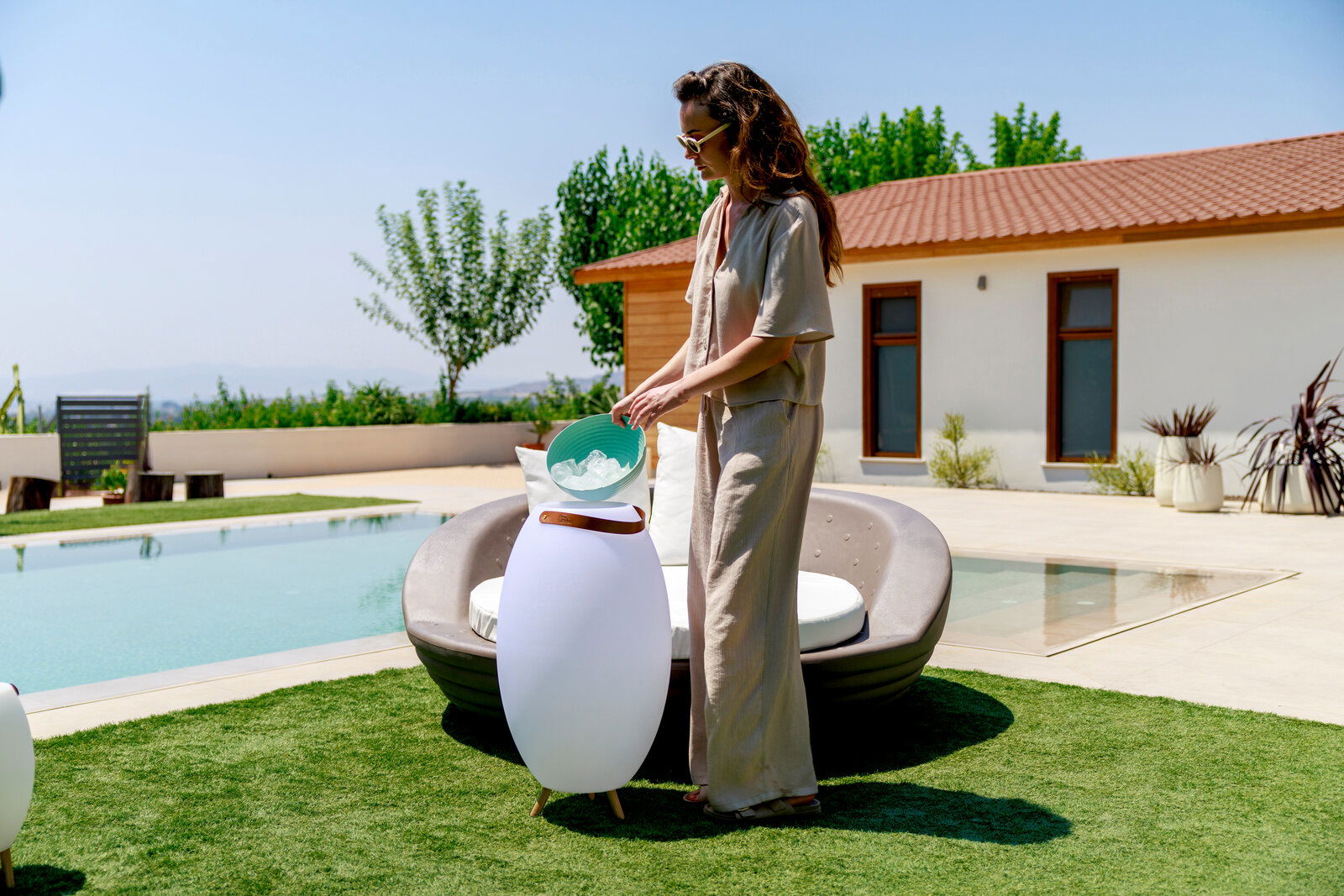 Frau füllt Eis aus einer Schüssel in einen weißen, eiförmigen Outdoor-Kühler mit Holzgriff neben einem runden Daybed am Pool; Seitenansicht in leichter Untersicht.