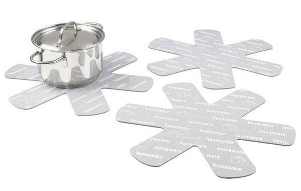 Drei graue Pfannenschützer in sternförmigem Design, auf einer weißen Oberfläche liegend, mit einem Edelstahltopf im Hintergrund. Perspektive von oben.