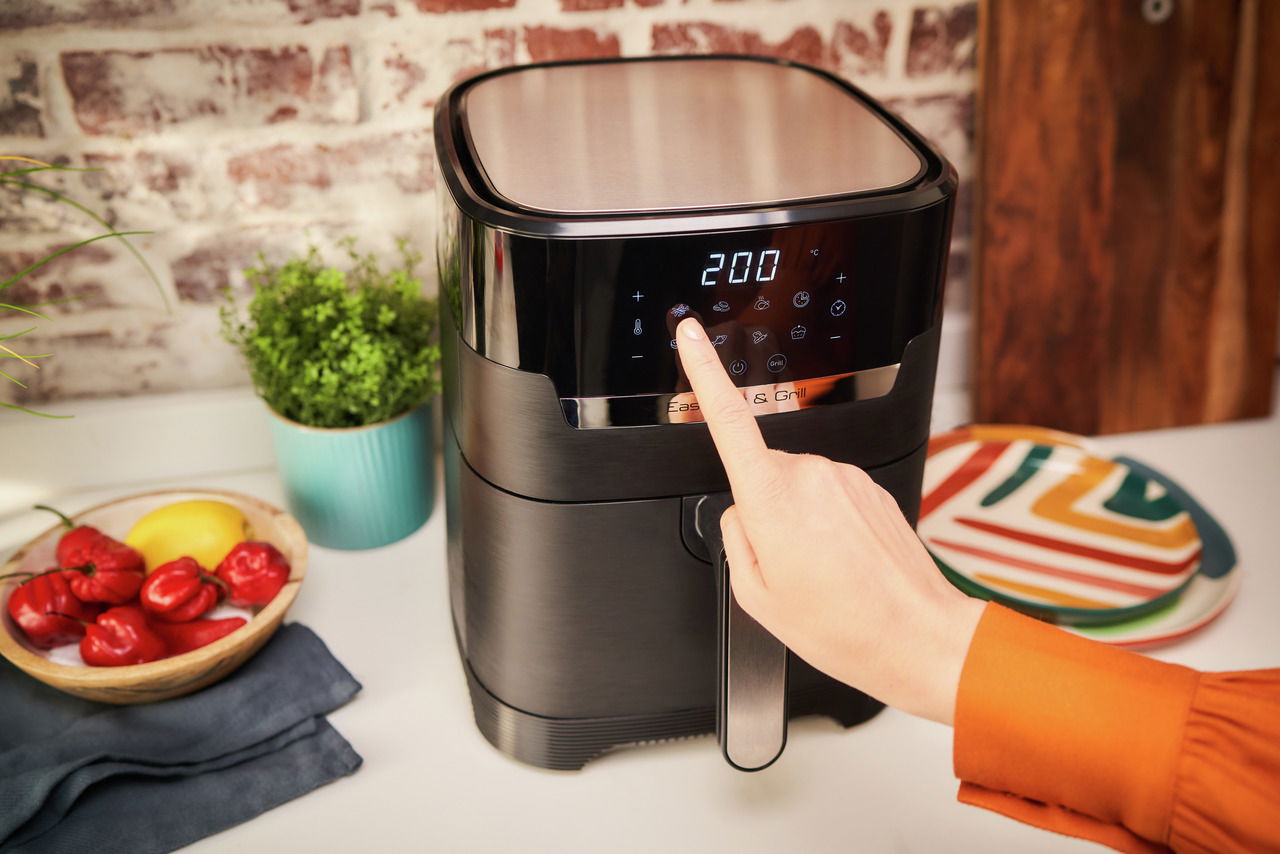 Nahaufnahme einer schwarzen Heißluftfritteuse mit digitalem Display, auf dem 200 Grad angezeigt werden. Eine Hand bedient das Touch-Panel. Im Hintergrund sind Küchenutensilien und Dekorationen zu sehen.