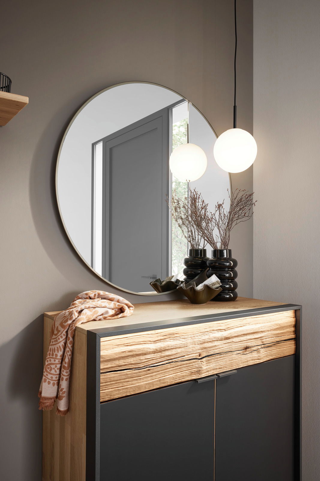 Runder Spiegel mit einem Durchmesser von 95 cm, an einer Wand über einer Kommode mit Holz- und Metallelementen angebracht. Im Spiegel spiegelt sich eine moderne Hängelampe wider. Perspektive: Frontalansicht.