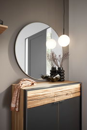 Runder Spiegel mit einem Durchmesser von 95 cm, an einer Wand über einer Kommode mit Holz- und Metallelementen angebracht. Im Spiegel spiegelt sich eine moderne Hängelampe wider. Perspektive: Frontalansicht.