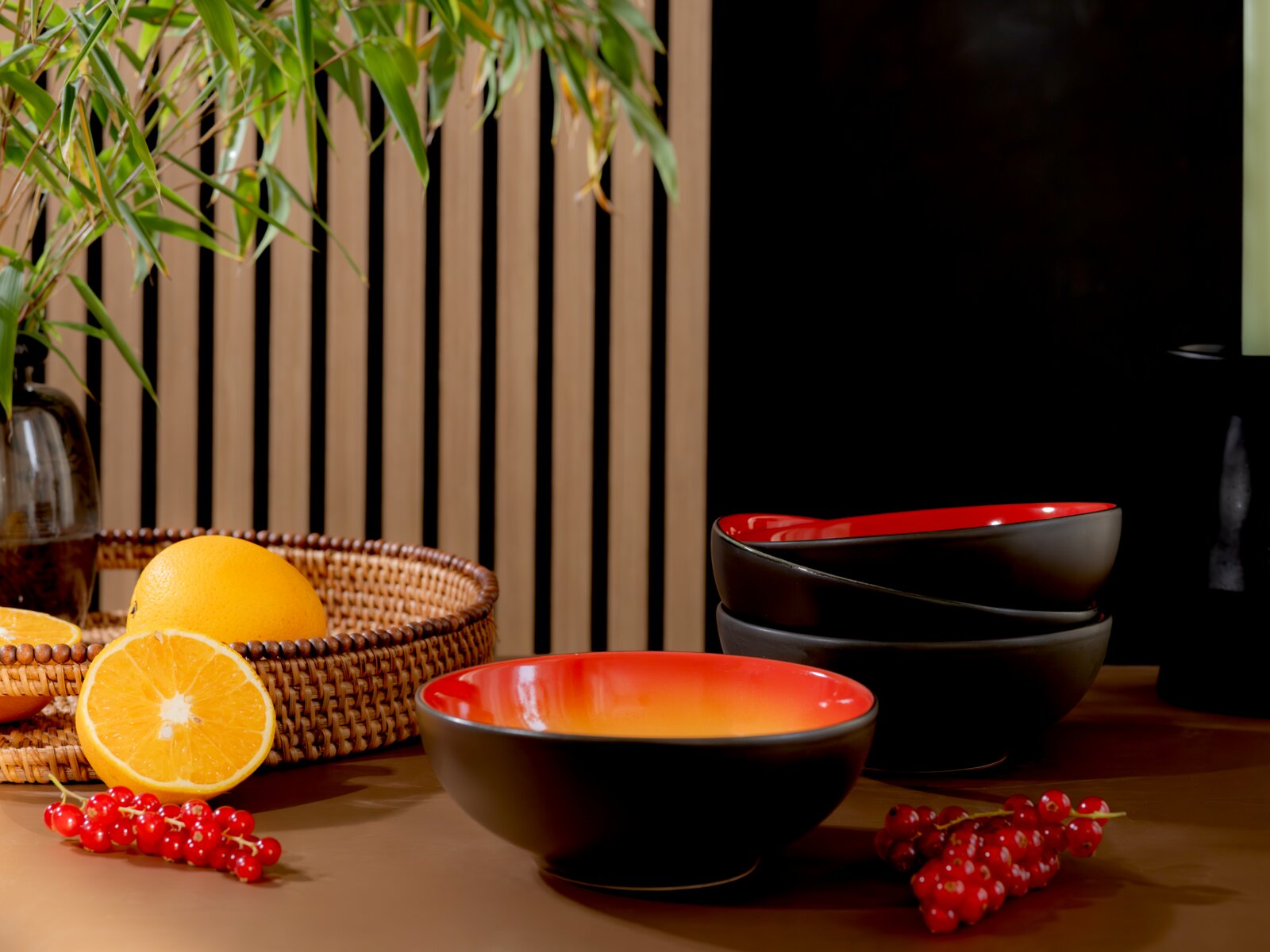 Drei schwarze Keramikschalen mit leuchtend roter Innenglasur, aus leicht erhöhter Frontalperspektive neben Orangen und roten Johannisbeeren arrangiert