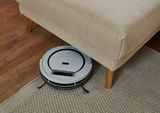 Ein Saugroboter reinigt den Boden unter einem beigen Longchair aus der Perspektive von oben.