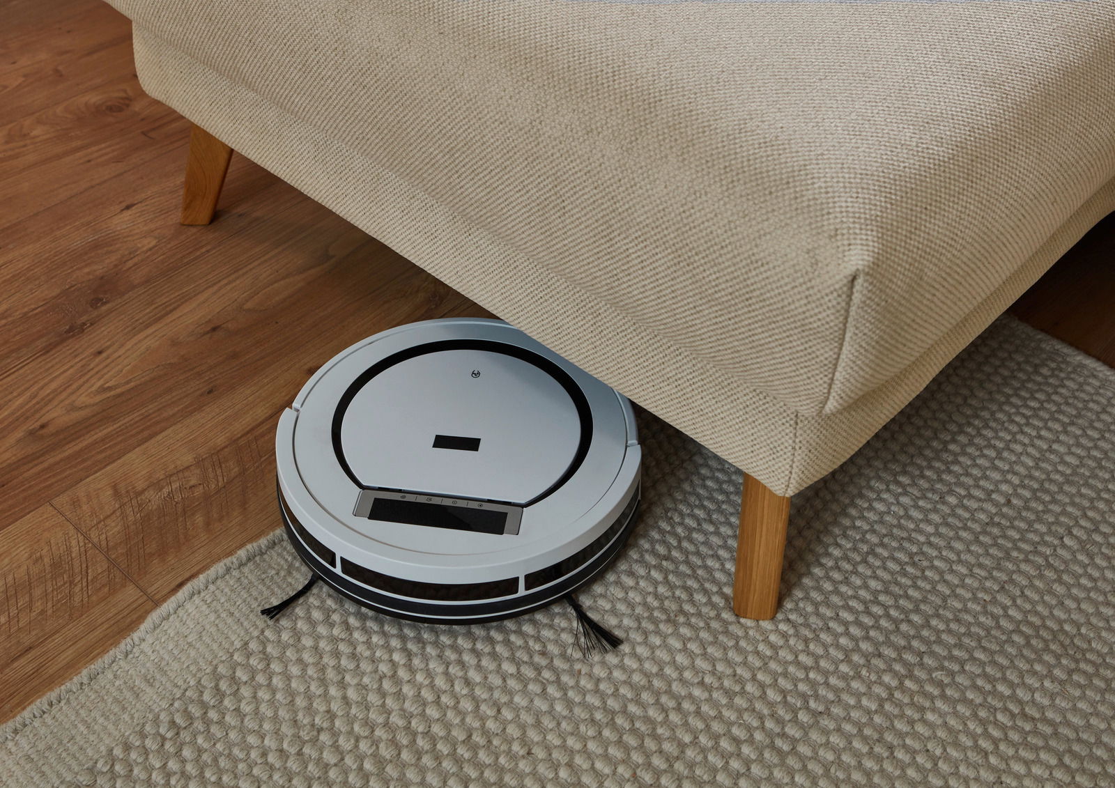 Ein Saugroboter reinigt den Boden unter einem beigen Longchair aus der Perspektive von oben.