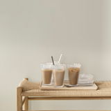 Drei Pilastro Trinkgläser mit Getränken und Löffeln, auf einem Tablett auf einem geflochtenen Tisch, seitliche Perspektive.