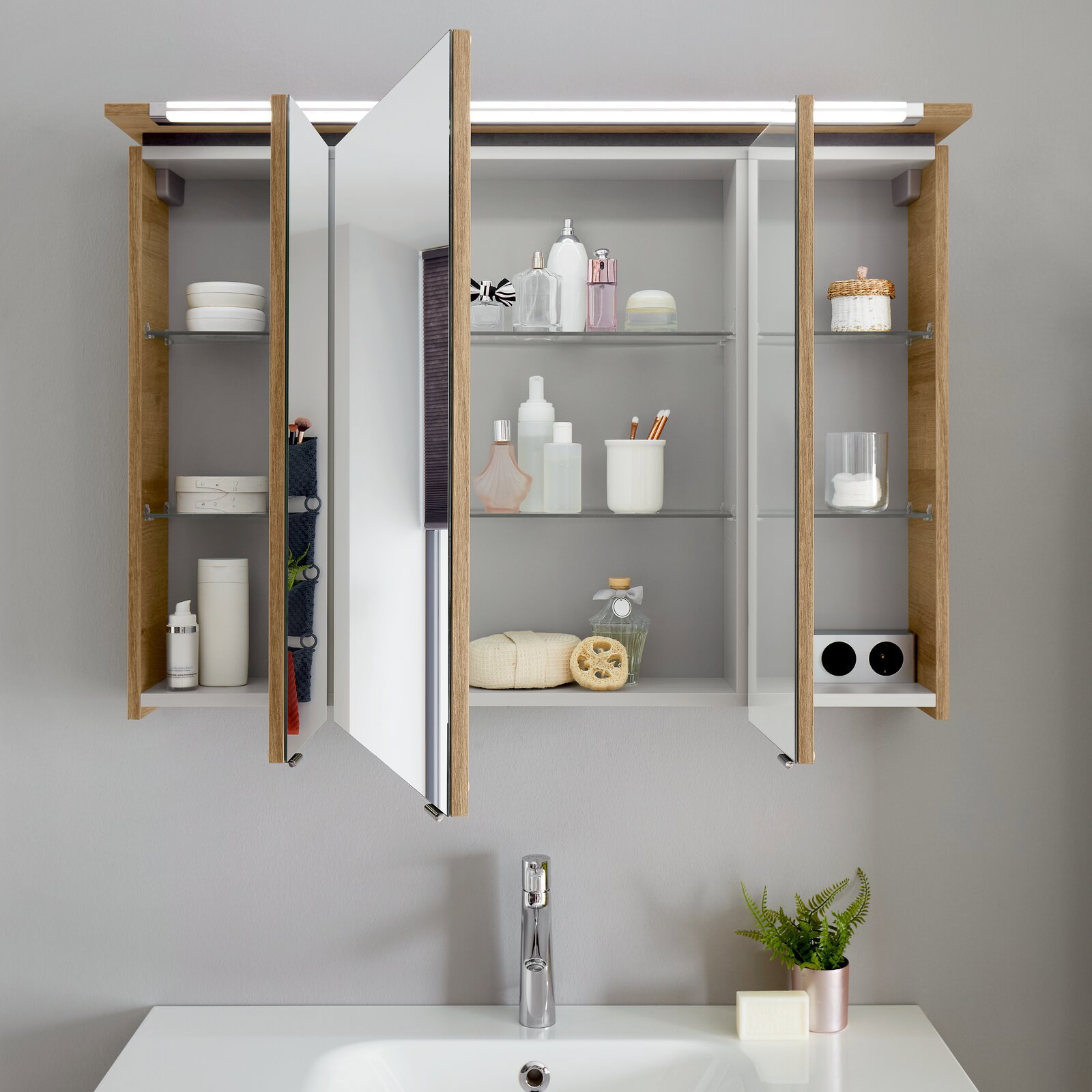 Badezimmerschrank mit Spiegel und Holzrahmen, geöffnet, mit verschiedenen Pflegeprodukten und Accessoires auf den Regalen, aus frontaler Perspektive.