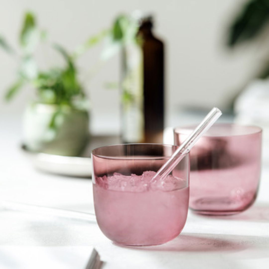 Zwei rosafarbene Wassergläser mit Eiswürfeln und einem Strohhalm, auf einem Tisch mit Pflanzen im Hintergrund, aus der Seitenperspektive fotografiert.