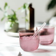 Zwei rosafarbene Wassergläser mit Eiswürfeln und einem Strohhalm, auf einem Tisch mit Pflanzen im Hintergrund, aus der Seitenperspektive fotografiert.