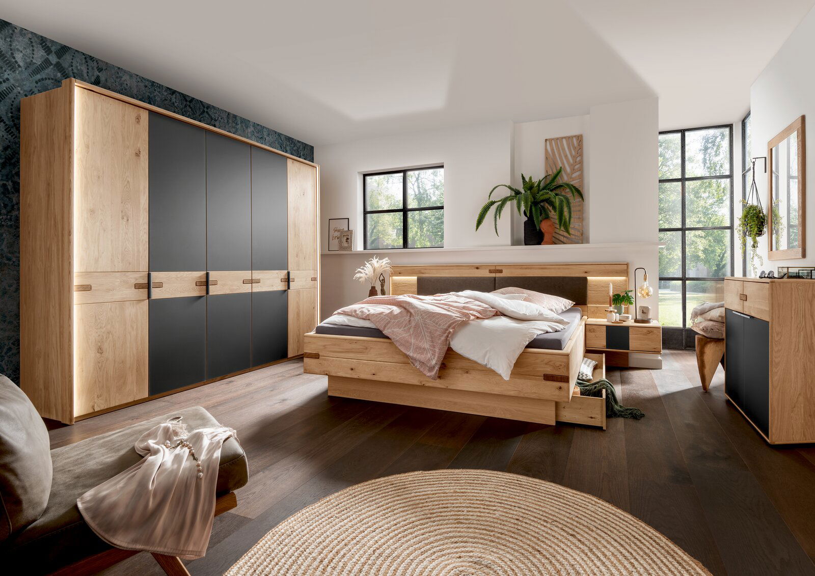 Linea Natura Nachtkonsole links TABILO Modernes Schlafzimmer mit Holz- und Schwarzakzenten, bestehend aus einem großen Kleiderschrank, einem Bett mit integrierten Schubladen und zwei Nachttischen, aus der Perspektive von der Zimmerecke aus gesehen.