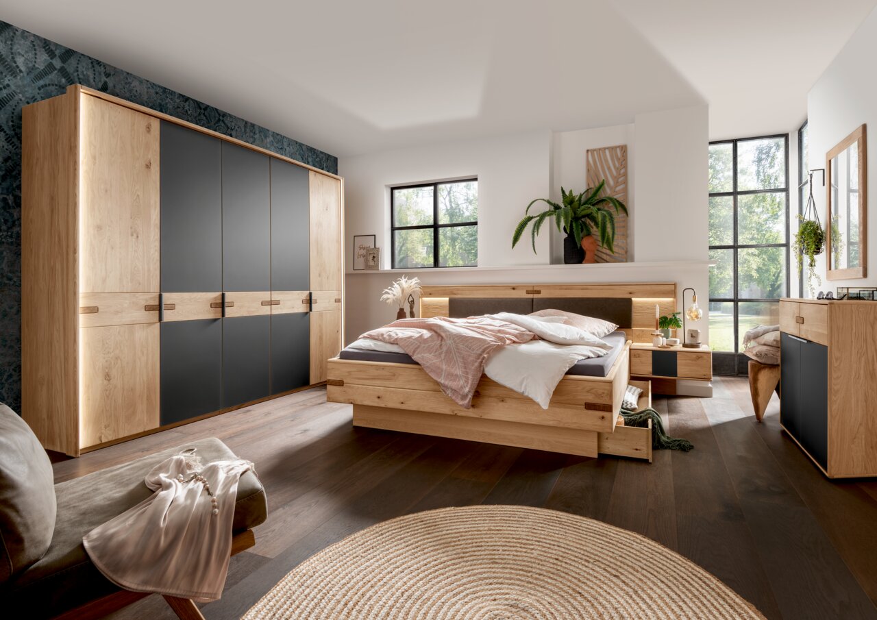 Linea Natura Nachtkonsole links TABILO Modernes Schlafzimmer mit Holz- und Schwarzakzenten, bestehend aus einem großen Kleiderschrank, einem Bett mit integrierten Schubladen und zwei Nachttischen, aus der Perspektive von der Zimmerecke aus gesehen.
