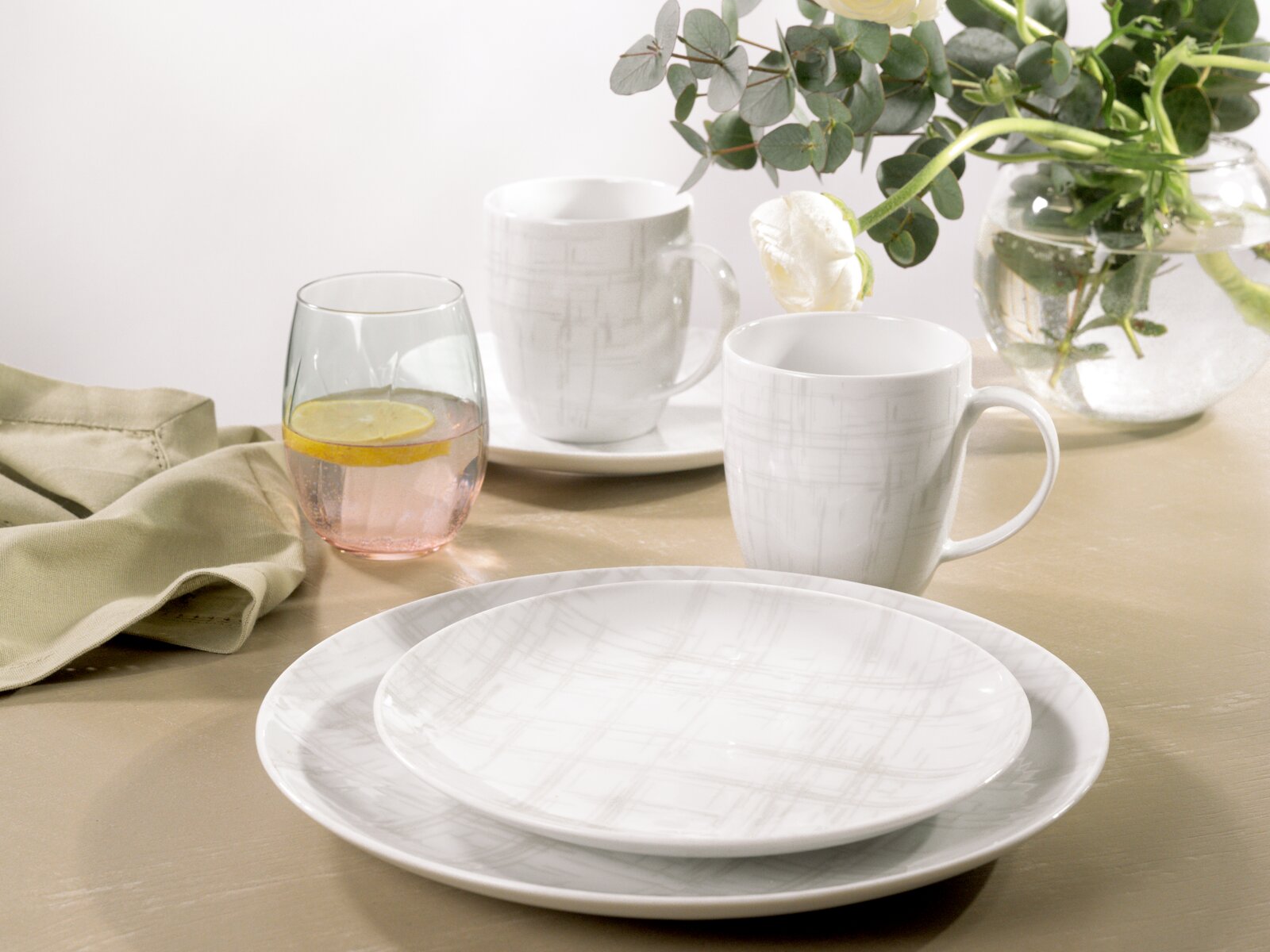 Weißes Geschirr-Set mit dezentem grauen Linienmuster (Teller und Tassen) auf gedecktem Tisch, fotografiert in leicht schräger Perspektive von vorne/oben, mit Glas und Blumen im Hintergrund