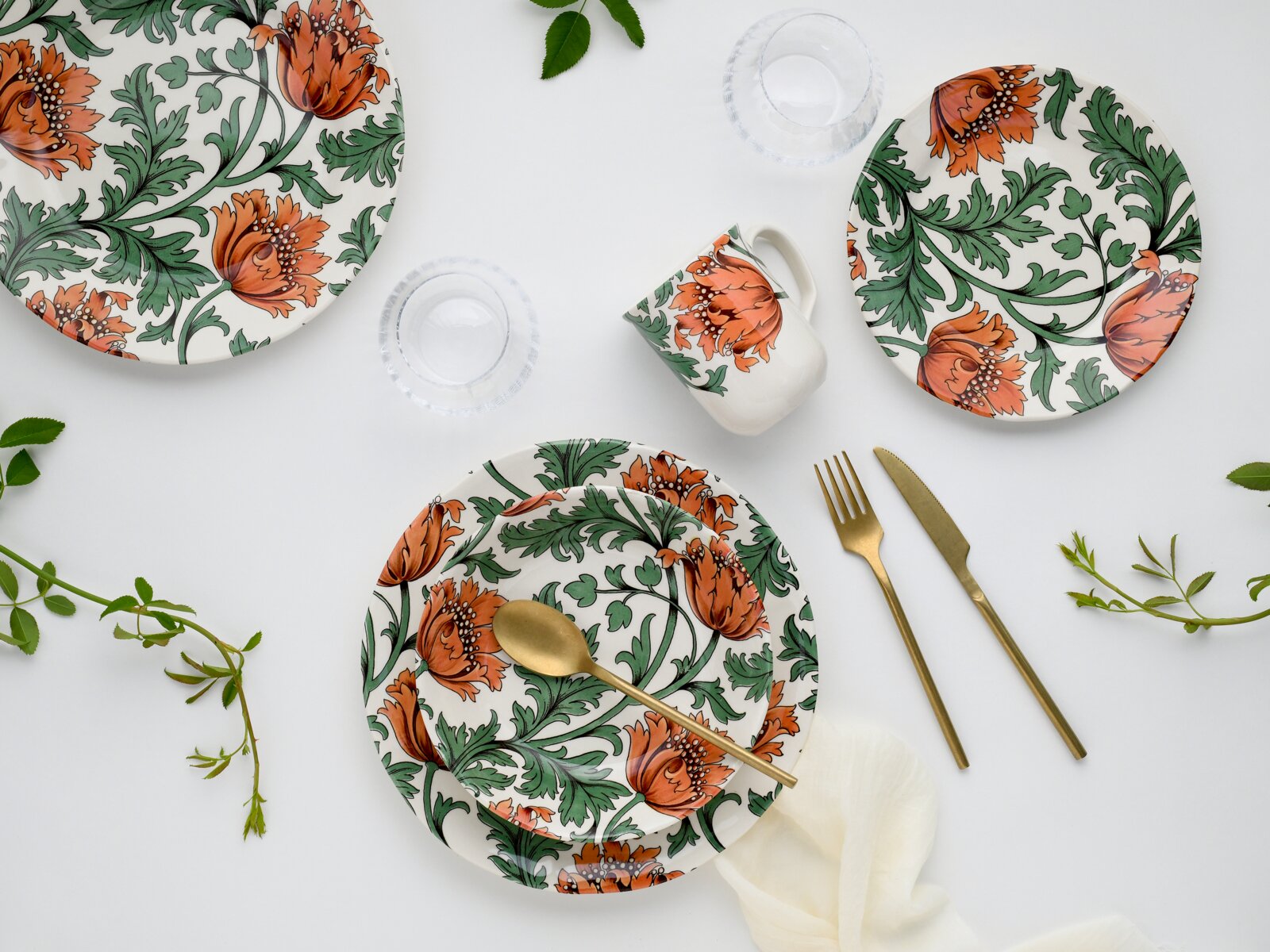 Draufsicht auf ein stilvolles Tischgedeck mit weißem Porzellan-Geschirr im orange-grünen Blumenmuster, Tasse und Tellern, dazu goldfarbenes Besteck und Wassergläser auf hellem Hintergrund.