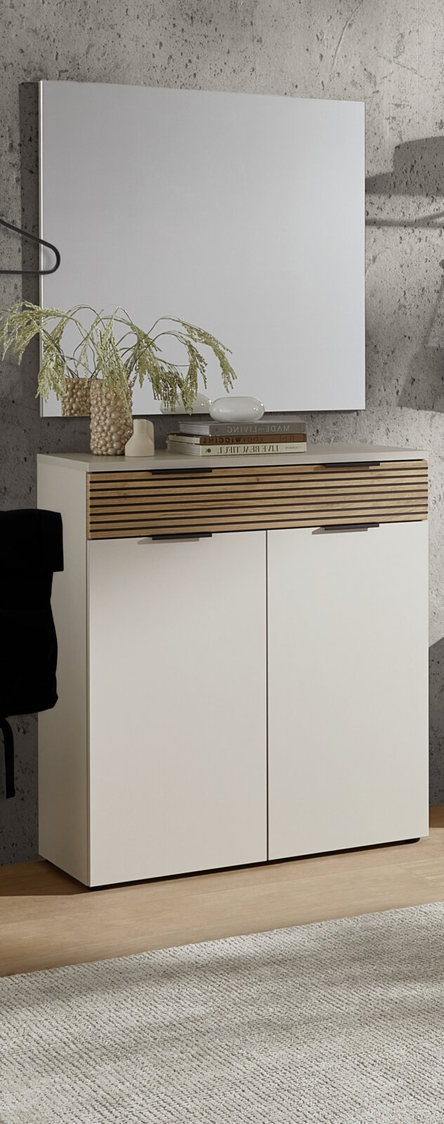 Voleo Garderoben-Set INDO Modernes Sideboard/Kommode in Weiß mit horizontaler Holzlamellen-Blende und zwei Türen, frontale Ansicht leicht von links, vor Betonwand mit Spiegel darüber; dekoriert mit Vase, Büchern und Schale.