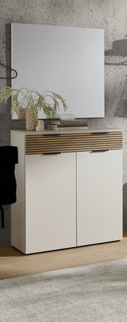 Modernes Sideboard/Kommode in Weiß mit horizontaler Holzlamellen-Blende und zwei Türen, frontale Ansicht leicht von links, vor Betonwand mit Spiegel darüber; dekoriert mit Vase, Büchern und Schale.