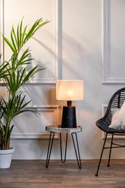 Moderner Lampenschirm auf einem kleinen Tisch, seitliche Perspektive, neben einer Pflanze und einem Stuhl.