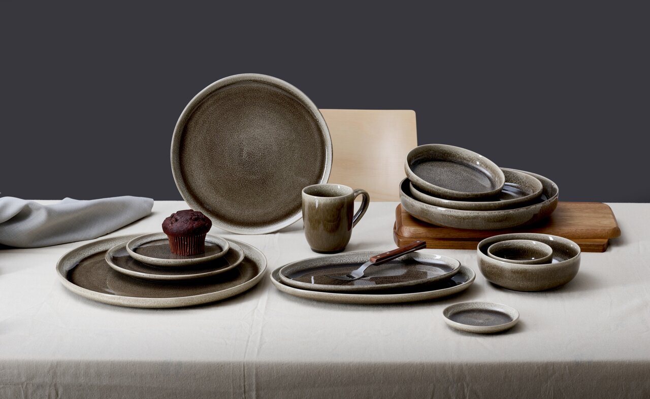 Vielseitiges Geschirrset in Erdtönen, bestehend aus Tellern, Schalen und einer Tasse, auf einem Tisch arrangiert. Perspektive von vorne.