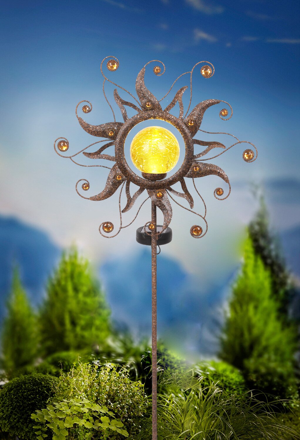 Decoratieve zonne-lamp in de vorm van een zon met een gele lamp, gemonteerd op een stok, tegen een achtergrond van groene planten en een blauwe lucht, zijaanzicht.