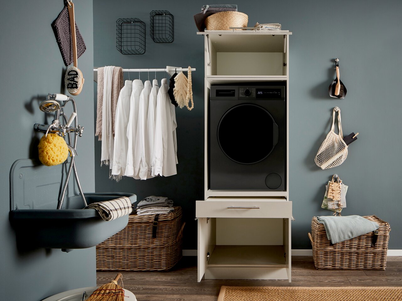 Frontalansicht eines modernen Waschturms: schwarze Frontlader-Waschmaschine in einem weißen Schrank mit offener Schublade, daneben Wäschekorb, Hemden auf Kleiderstange und Badaccessoires an grauer Wand.
