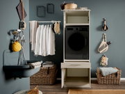 Frontalansicht eines modernen Waschturms: schwarze Frontlader-Waschmaschine in einem weißen Schrank mit offener Schublade, daneben Wäschekorb, Hemden auf Kleiderstange und Badaccessoires an grauer Wand.