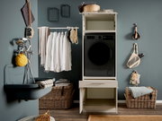 Frontalansicht eines modernen Waschturms: schwarze Frontlader-Waschmaschine in einem weißen Schrank mit offener Schublade, daneben Wäschekorb, Hemden auf Kleiderstange und Badaccessoires an grauer Wand.