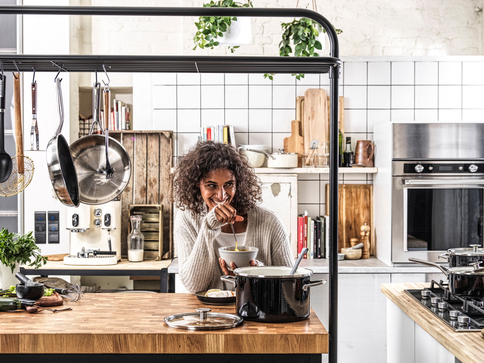 KitchenAid Topfset 4-tlg. COOKWARE Frau in moderner Küche mit einem schwarzen Topfset auf dem Herd, fröhlich kochend. Perspektive: Frontalansicht.